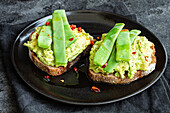 Appetizing toasts with fresh guacamole and green peas pods garnished with red peppers and served on black plate