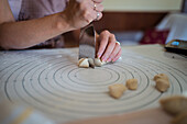High angle of crop anonymous lady cutting raw dough with putty knife while preparing homemade dumplings sitting at table in kitchen