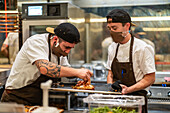 Male cook adding filling on flatbread on stainless tray in hands on colleague wearing protective mask and uniform in restaurant