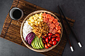 Draufsicht auf eine appetitliche bunte Poke-Schale mit frischem Lachs und Reis, serviert mit reifen Kirschtomaten, Avocadoscheiben, Zwiebeln und Mango auf einem Bambustablett mit Sojasauce