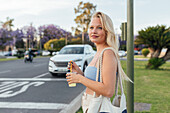 Side view of cheerful female standing with cold lemonade in plastic cup in street in summer