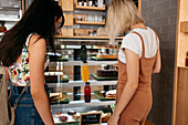 Back view anonymous female interacting with partner while showing delicious cakes behind showcase in pastry shop