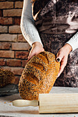 Anonyme Bäckerin hält Brot mit Sonnenblumenkernen auf einem Tisch in einer Backstube