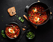 Top view of chakchouka with sunny side up egg in delicious tomato sauce with rye bread piece in bowl between fresh cilantro on dark background