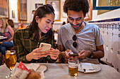 Happy young multiracial couple in casual clothes using smartphones while sitting together at table with glasses of beer in modern restaurant