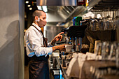 Side view of waiter in uniform and protective mask pressing buttons on coffee machine while brewing beverage in restaurant during coronavirus pandemic