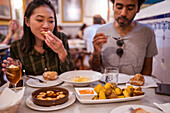 Crop hungry multiracial couple with dark hair in casual clothes eating appetizing dishes in modern restaurant during dinner