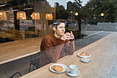 Junger Mann sitzt am Holztresen in der Nähe des Fensters und schaut nachdenklich weg, während er einen aromatischen Kaffee und ein Croissant in einer modernen Cafeteria genießt