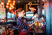 Seitenansicht einer Barkeeperin bei der Auswahl eines alkoholischen Getränks in einer Flasche für die Zubereitung eines Cocktails in einer Bar