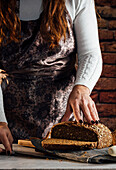 Crop anonymous female baker with knife cutting loaf of fresh bread with sunflower seeds on table in bakehouse