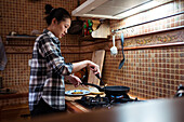 Side view of focused young Asian female in casual clothes cooking yummy pancakes using spatula while standing in kitchen