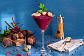Glass goblet of tasty beetroot cream garnished with quail eggs and parsley placed near napkin with spoon and fresh ingredients against blue background