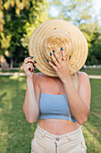 Anonymous female covering face with straw hat while standing in park in summer