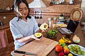 Young Asian woman halving ripe avocado over cutting board while cooking healthy salad following online recipe on laptop with black screen in kitchen at home