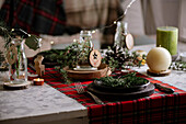 Christmas table setting with wreath on the plate, decorative wooden ornaments and red checkered tablecloth with yellow lights on the background