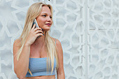 Ruhig lächelnde Frau mit blondem Haar und in Sommerkleidung steht in der Stadt und spricht mit dem Handy, während sie wegschaut