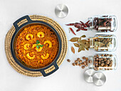 Leckere Reis-Paella mit Garnelen im Topf auf Korbständer und Glasflaschen mit rotem Pfeffer, Lorbeerblättern und Nüssen von oben