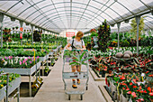 Young female customer in protective mask for coronavirus prevention with shopping trolley doing purchases in gardening market and choosing houseplants inside of glasshouse
