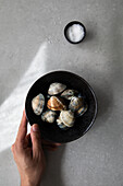 Top view of cropped unrecognizable person holding bowl with uncooked clams and salt placed on gray tabletop during food preparation
