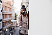Ethnische Frau in gestreifter Kleidung mit professionellem Fotogerät schaut tagsüber auf dem Balkon in die Kamera