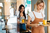 Young content girlfriends standing with delicious food and refreshing drinks in cafeteria