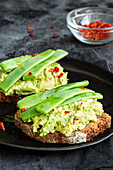 Appetizing toasts with fresh guacamole and green peas pods garnished with red peppers and served on black plate