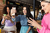 Smiling young female friends wearing casual clothes taking selfie and browsing mobile phones while gathering for lunch in restaurant