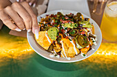 Blick von oben auf eine nicht erkennbare weibliche Person, die köstliche traditionelle mexikanische Nachos mit Jalapeno-Chili-Paprika, Käse und Guacamole-Sauce am Tisch in einem Restaurant isst
