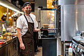Seitenansicht eines männlichen Kochs in Uniform und Schutzmaske, der in der Küche eines Restaurants während einer Coronavirus-Pandemie ein Gericht im Ofen zubereitet