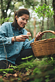 From below female in eyeglasses picking edible Ramaria mushroom from ground covered with fallen dry leaves and putting into wicker basket