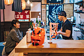 Seitenansicht einer asiatischen Frau in Freizeitkleidung, die am Tresen sitzt und sich mit einem männlichen Angestellten einer modernen Ramen-Bar unterhält