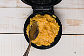 Top view of sweet pumpkin batter in black metal cheese waffle iron with spoon placed on wooden table in light kitchen