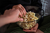 Cropped unrecognizable couple wearing casual clothes sitting on couch and eating popcorn at home