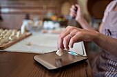 Crop anonymous female cook weighing dough on electronic scale while preparing jiaozi sitting at table in kitchen