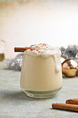 Glas Milchpunsch mit Zimtpulver auf geschlagenem Eiweiß vor Tannenzapfen am Weihnachtstag auf hellem Hintergrund
