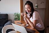 Fokussierte Asiatin mit heißem Getränk surft auf ihrem Netbook, während sie beim Frühstück in einer modernen Küche zu Hause am Tresen sitzt