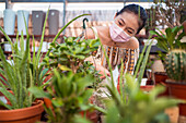 Young ethnic female buyer in disposable mask choosing potted plants while looking away in garden shop