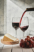 Semi sweet red wine pouring into wineglass placed on wooden table near triangle cheese piece and grapes in studio