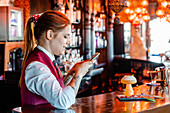 Side view of cheerful female bartender taking photo of sour cocktail in glass served on counter in pub