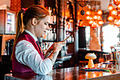 Seitenansicht einer fröhlichen Barkeeperin, die einen sauren Cocktail im Glas auf dem Tresen eines Pubs fotografiert