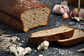 Loaf and slices of healthy bread placed on table with scattered sesame seeds and garlic cloves in kitchen