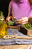 Crop unrecognizable woman in purple sweater cutting herbs twigs with scissors at table with essential oil glass bottles and rope with small chest on textile