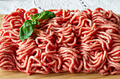 From above of fresh raw mixed pork and beef minced meat on wooden cutting board with green basil leaves placed on table during cooking process