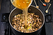 From above of broth pouring into metal pot with mushrooms on modern induction cooker while cooking soup near various spices
