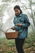 Serious female in eyeglasses carrying wicker basket and picking edible mushrooms in woods in hazy weather