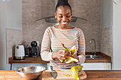 Zufriedene Afroamerikanerin in Kleidung mit gestreiftem Ornament schält Kochbananen am Tisch im Haus