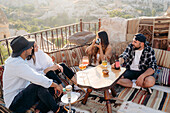 Von oben auf eine Frau, die ein junges Paar mit einer Sofortbildkamera fotografiert, während sie in einer Bar mit Freunden auf einer Terrasse in Kappadokien, Türkei, Cocktails trinkt