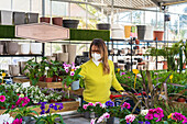 Female customer in protective mask standing with shopping cart in garden center and picking blooming potted plants