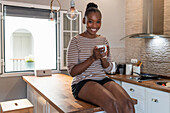 Happy young dreamy African American female with mug of hot drink sitting on table and looking at camera indoors