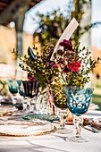 Nahaufnahme einer festlich gedeckten Tafel mit Kristallgläsern, Besteck und Serviette auf einem Teller neben einem Strauß frischer Blumen für eine Hochzeit